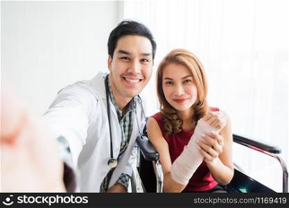 Selfie of smiling man doctor with a female patient wear arm splint for better healing sit in a wheelchair using a smartphone In the room hospital background.