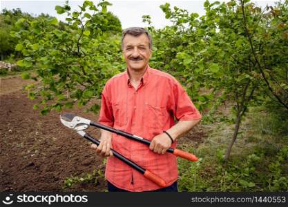 Senior adult caucasian man Standing holding telescopic branches Pruning shears for trimming Gardening squid scissors in the farm on the plantation at countryside in day front view wearing red shirt