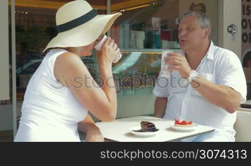 Senior adult wife and husband drinking coffee at table in cafe, then taking selfie using selfie stick. Summertime, holiday.