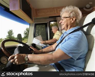 Senior adult woman driving RV and smiling while man reads map in passenger seat.