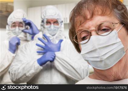 Senior Adult Woman Wearing Medical Face Mask In Doctor Office With Nurses In HAZMAT Suits Behind.