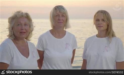 Senior and young women in white t-shirts wearing pink breast cancer awareness ribbons, outdoor shot on sea background. Support for people suffering from disease