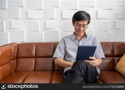 Senior Asian man playing tablet on sofa in living room at home Portrait of Asian elderly man is Relaxing and Happiness With playing tablet