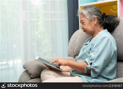 Senior asian woman wearing glasses and relaxing at home on a sofa and using tablet for reading news and e-book. Concept of technology for elderly.