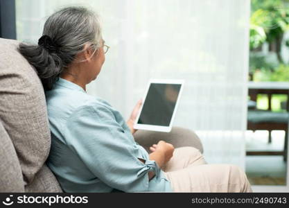 Senior asian woman wearing glasses and relaxing at home on a sofa and using tablet for reading news and e-book. Concept of technology for elderly.