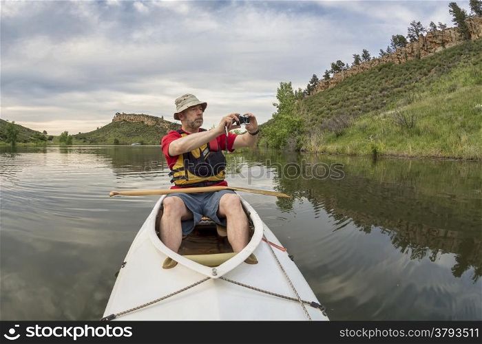 senior athletic paddler in a decked expedition canoe photographing on a lake with green vegetation - Horsetooth Reservoir, Fort Collins, Colorado