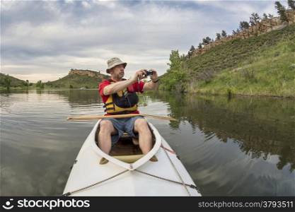 senior athletic paddler in a decked expedition canoe photographing on a lake with green vegetation - Horsetooth Reservoir, Fort Collins, Colorado