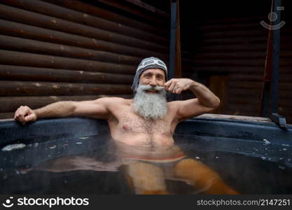 Senior bearded man bathing in steaming outside vat with warm water looks at camera closeup portrait. Senior bearded man in outside vat portrait