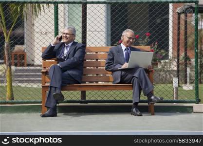 Senior businessman on phone call with other executive using laptop while sitting on bench in a tennis court
