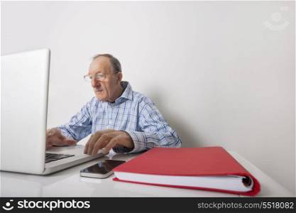 Senior businessman using laptop with book binder and cell phone on office desk