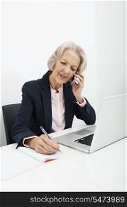 Senior businesswoman writing in book while answering cell phone at desk in office