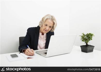 Senior businesswoman writing in book while using laptop at desk in office