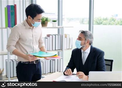 Senior Caucasian boss and young Asain businessman wearing mask preventing for Covid 19 virus holding folder work sheet and talking about job in office. Social distance and new normal lifestyle concept