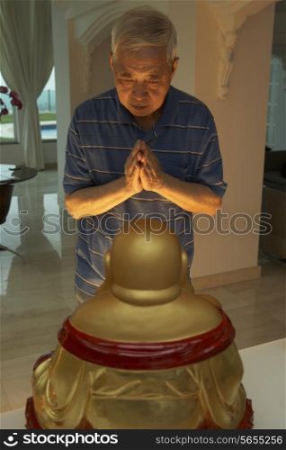 Senior Chinese Man Praying To Statue Of Buddha At Home