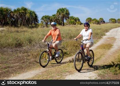 Senior couple bicycling at the beach, wearing safety helmets and sunglasses.