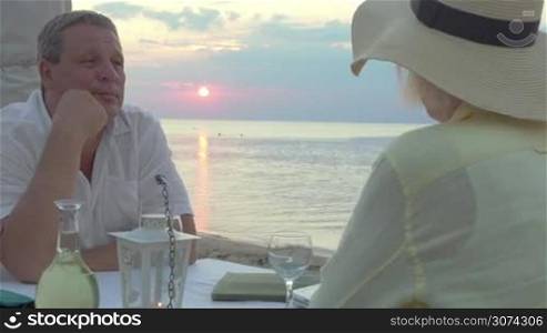 Senior couple having romantic evening outdoor. They sitting in the restaurant on the beach at sunset and drinking white wine