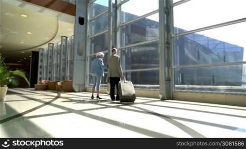 Senior couple of tourists walking with suitcase in airport and talking