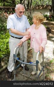 Senior couple outdoors. She&rsquo;s in a walker and he&rsquo;s helping her.