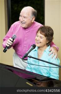 Senior couple performs. They both sing while she plays piano.