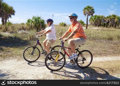 Senior couple riding bikes at the beach, wearing sunglasses and helmets. Focus on the woman.