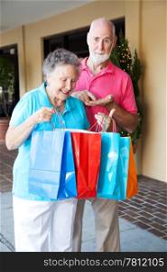 Senior couple shopping. She&rsquo;s happy with her purchases, and he&rsquo;s upset because she spent all his money.