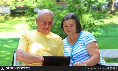 Senior couple sitting on a bench in the park and working on digital tablet pc
