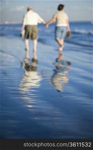 Senior couple walking on beach, reflecting in water