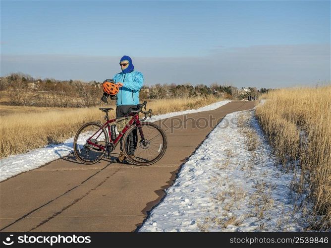 senior cyclist wearing warm balaclava is putting helmet on to ride a gravel bike on a biking trail on winter afternoon in Fort Collins, Colorado