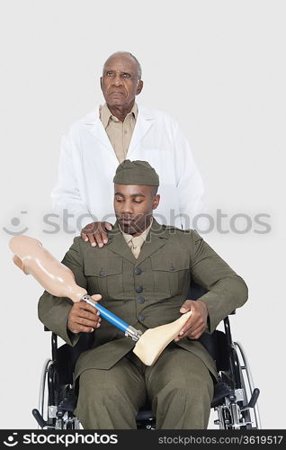 Senior doctor standing behind military officer holding artificial limb as he sits in wheelchair over gray background