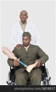 Senior doctor standing behind military officer holding artificial limb as he sits in wheelchair over gray background
