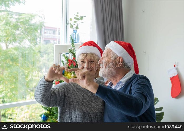 senior elderly caucasian old man and woman created heart shape sign with hand together in living room that decorated with christmas tree for christmas festival day, retirement lifestyle concept
