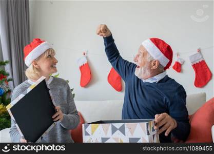 senior elderly caucasian old man and woman help open big gift presents box and happy together in living room that decorated with christmas tree for christmas festival day, lifestyle concept
