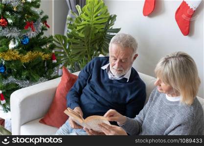 senior elderly caucasian old man and woman reading book and sitting on sofa to discussion together in living room that decorated for christmas festival day in the morning, retirement family concep