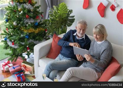 senior elderly caucasian old man and woman sitting on sofa, using and play internet tablet together in living room that decorated for christmas festival day in the morning, retirement family concept
