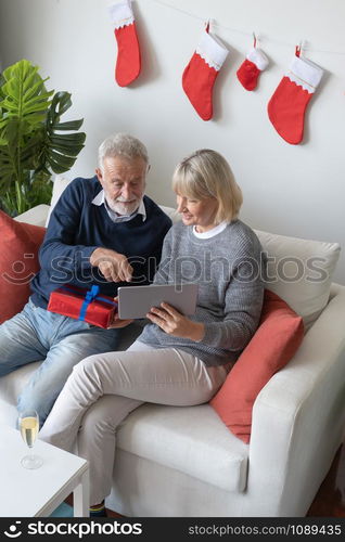 senior elderly caucasian old man and woman sitting on sofa, using and play internet tablet together in living room that decorated for christmas festival day in the morning, retirement family concept