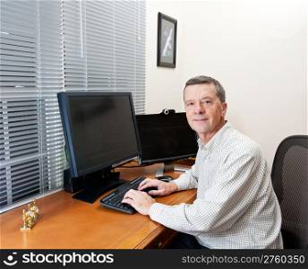 Senior executive in home office with two monitors and keyboard on leather desk and facing the camera