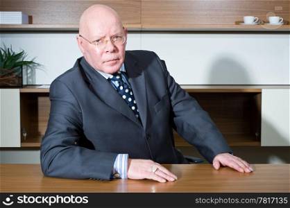 Senior executive posing in a modern, sparce, office, looking stern, and leaning on his desk