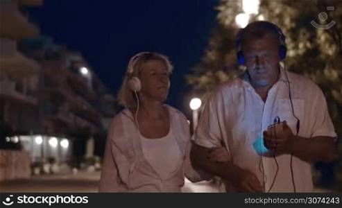 Senior family couple in headphones enjoying music while walking on resort at night. Relaxing together in quiet evening