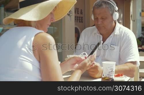 Senior family couple in street cafe on sunny summer day. Man in headphones listening to music or watching movie on pad while woman using smartphone