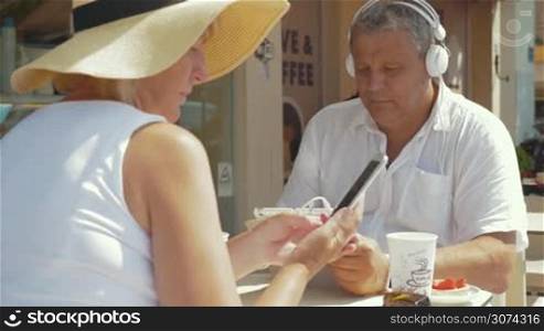 Senior family couple in street cafe on sunny summer day. Man in headphones listening to music or watching movie on pad while woman using smartphone