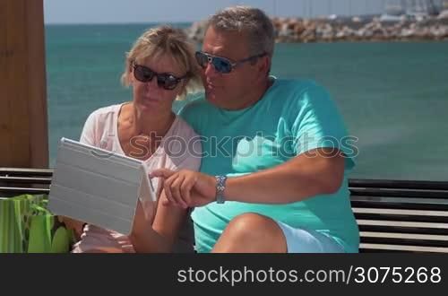 Senior family couple in sunglasses browsing the web on tablet computer while sitting on the beach on waterfront