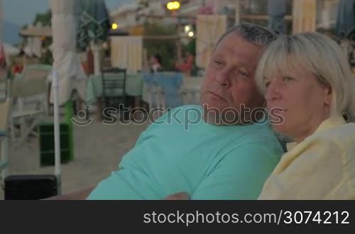 Senior family couple sitting on the beach, staring somewhere with relaxed look and talking. Outdoor cafe in background