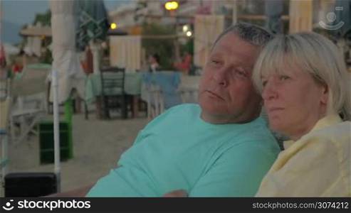 Senior family couple sitting on the beach, staring somewhere with relaxed look and talking. Outdoor cafe in background