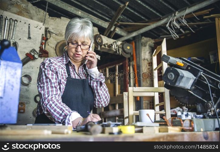 Senior female carpenter talking on the phone in her workshop. Female carpenter in her workshop