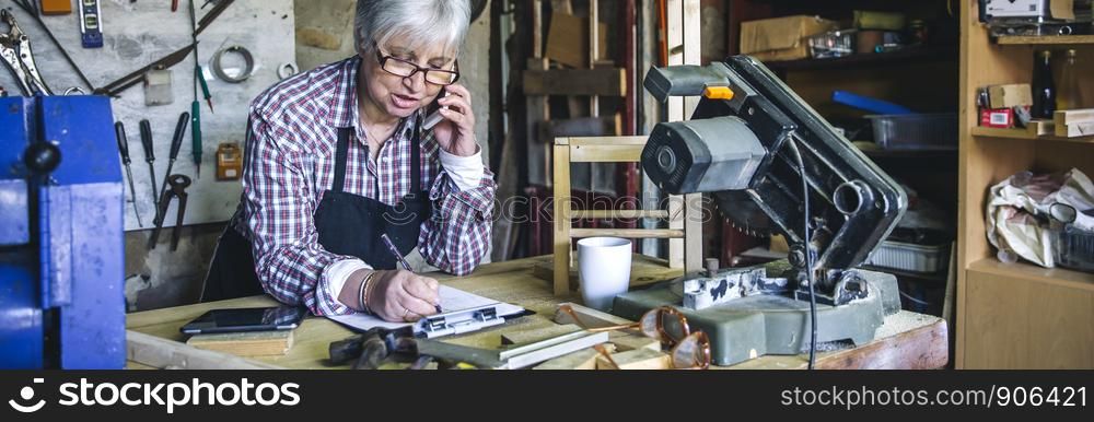Senior female carpenter talking on the phone in her workshop. Female carpenter in her workshop