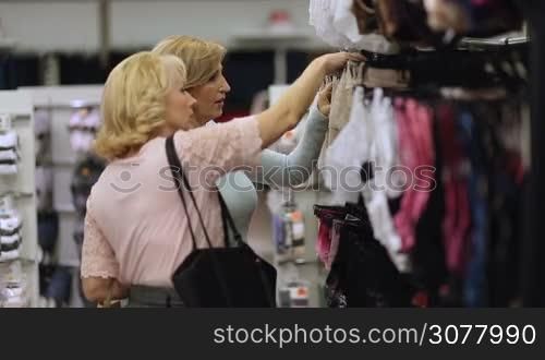 Senior female friends shopping and searching for clothes at clothing shop. Two shopaholic women in good mood choosing underpants, knickers and other lingerie while shopping in apparel boutique. Elegant mature ladies buying underwear at store.