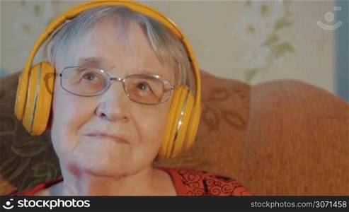 Senior gray-haired woman in yellow headphones sits on the sofa listening to music