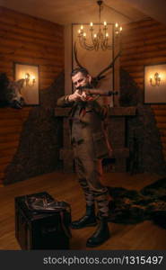 Senior hunter in retro traditional hunting clothing aims of the antique shotgun against old chest. Fireplace, stuffed wild animals, bear skin and other trophies on background
