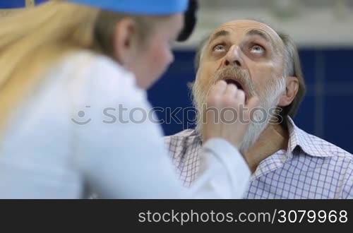Senior ill male patient checking his inflamed throat in medical office. Female ear nose and throat specialist examining elderly man patient using a tongue depressor and head mirror to look inside his mouth and tonsils at clinic.