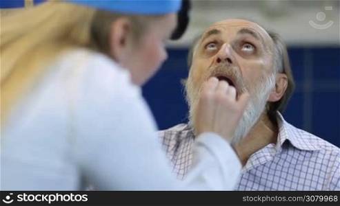 Senior ill male patient checking his inflamed throat in medical office. Female ear nose and throat specialist examining elderly man patient using a tongue depressor and head mirror to look inside his mouth and tonsils at clinic.